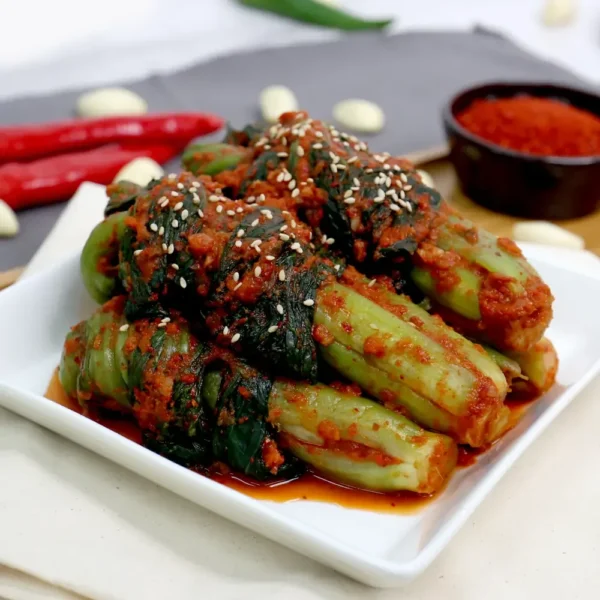 Close-up of HanMaru gat-kimchi (leaf-mustard kimchi) neatly tied and glazed with red seasoning, sprinkled with sesame seeds on a white plate.